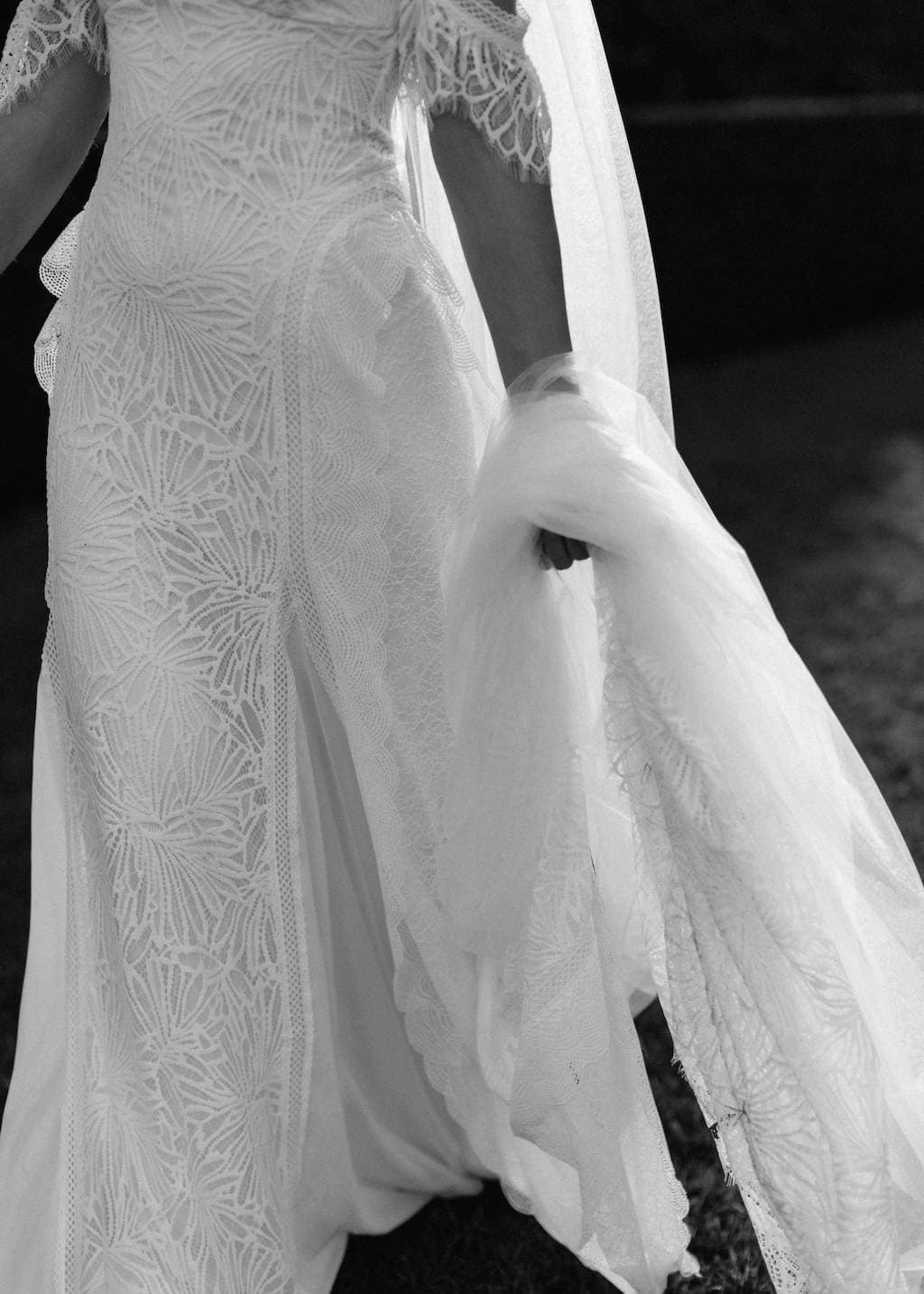 A black and white photo of a bride in a detailed, lace wedding dress. She is holding part of her long, flowing veil in her hand and walking outside, casting a shadow on the ground. The intricate pattern of the gown and veil is prominently displayed.