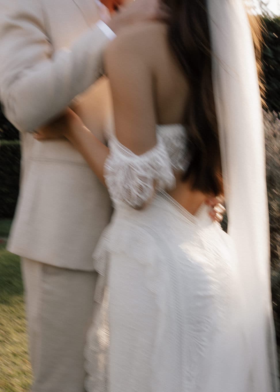 A blurred image of a bride and groom embracing outdoors. The bride is wearing a lace wedding dress and a veil, while the groom is dressed in a light-colored suit. They appear to be in a lush garden setting.