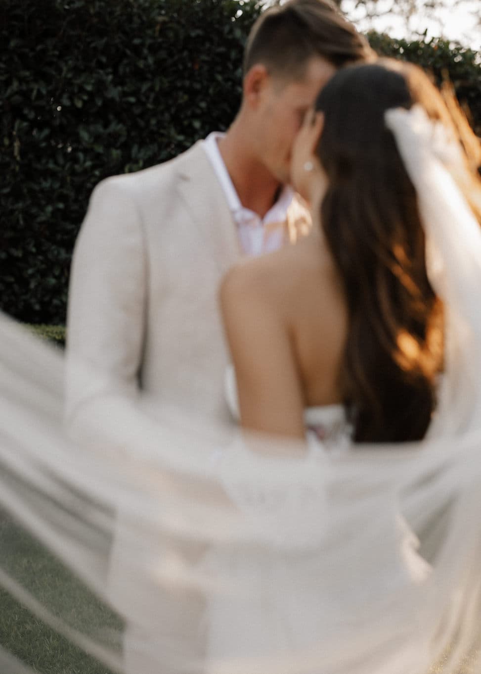 A blurred, romantic image of a bride and groom kissing outdoors. The bride, with long hair and a flowing veil, is facing the groom, who is wearing a light-colored suit. The background features green foliage.