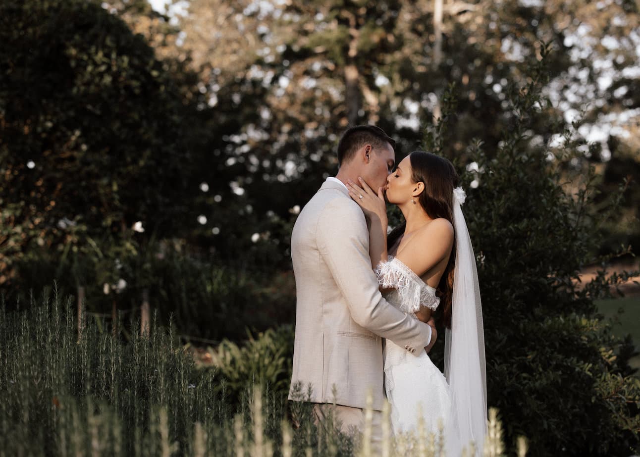 A couple dressed in wedding attire shares a kiss outdoors. The bride, in a white dress with a long veil, gently holds the groom's face. The groom, in a light-colored suit, embraces the bride. They are surrounded by greenery and trees, creating a romantic backdrop.