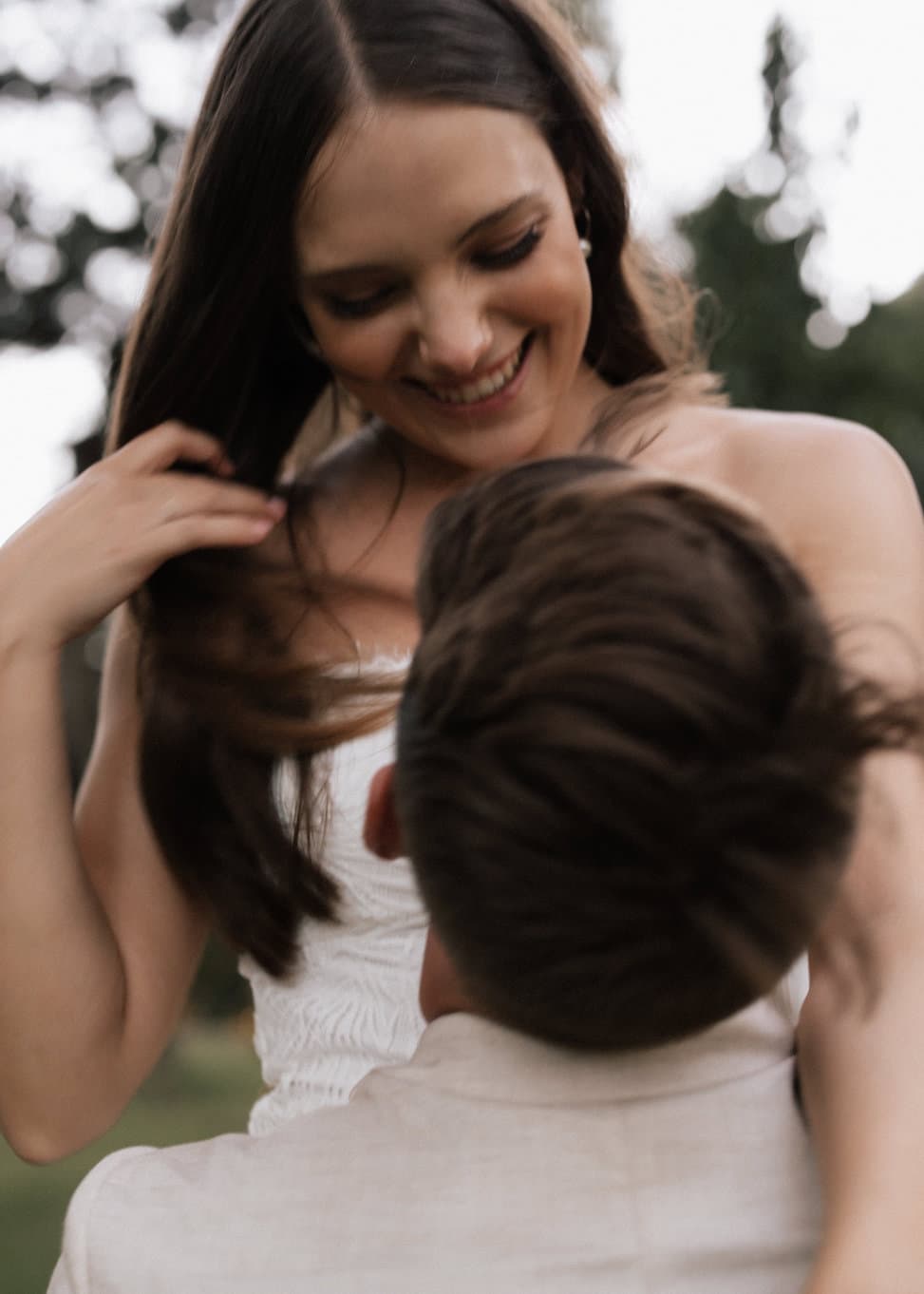 A woman with long brown hair smiles down at a man whose back is facing the camera. She gently touches her hair while wearing a white dress, and he is in a light-colored jacket. Blurry green foliage is in the background, suggesting an outdoor setting.