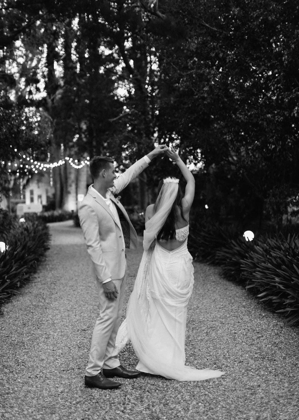 A couple dressed in wedding attire dances on a gravel path under string lights. The groom, in a light-colored suit, twirls the bride, who wears a long veil and flowing gown. They are surrounded by lush greenery in an outdoor setting, creating a romantic ambiance.