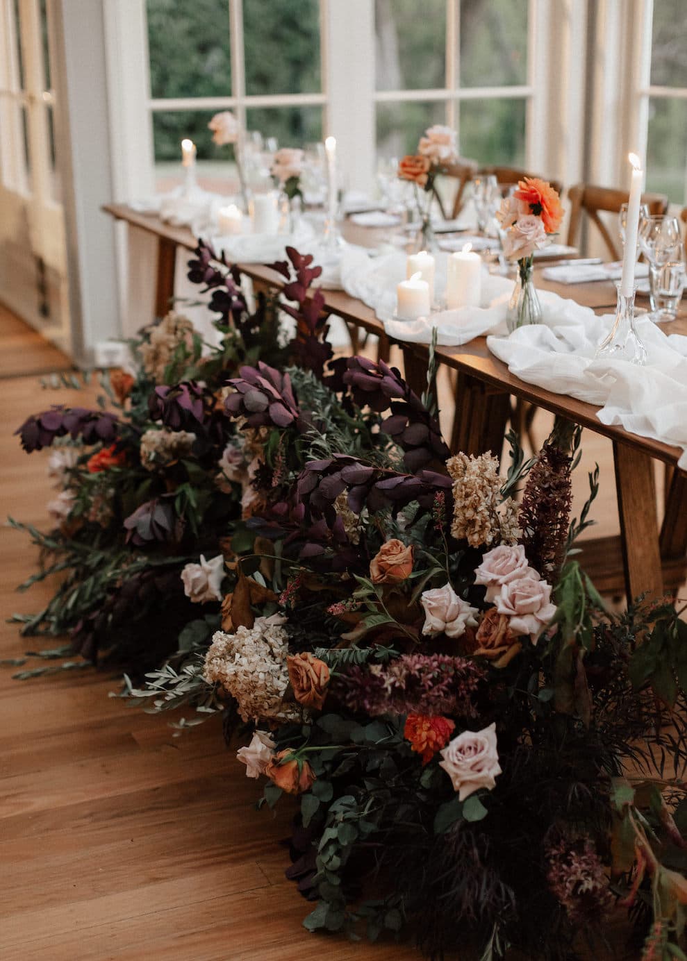 A beautifully decorated wooden table setup for an event. The table is adorned with white tablecloths, scattered flower arrangements in warm, muted tones, and lit candles. A lush floral arrangement with rich burgundy, peach, and cream flowers lines the base of the table.