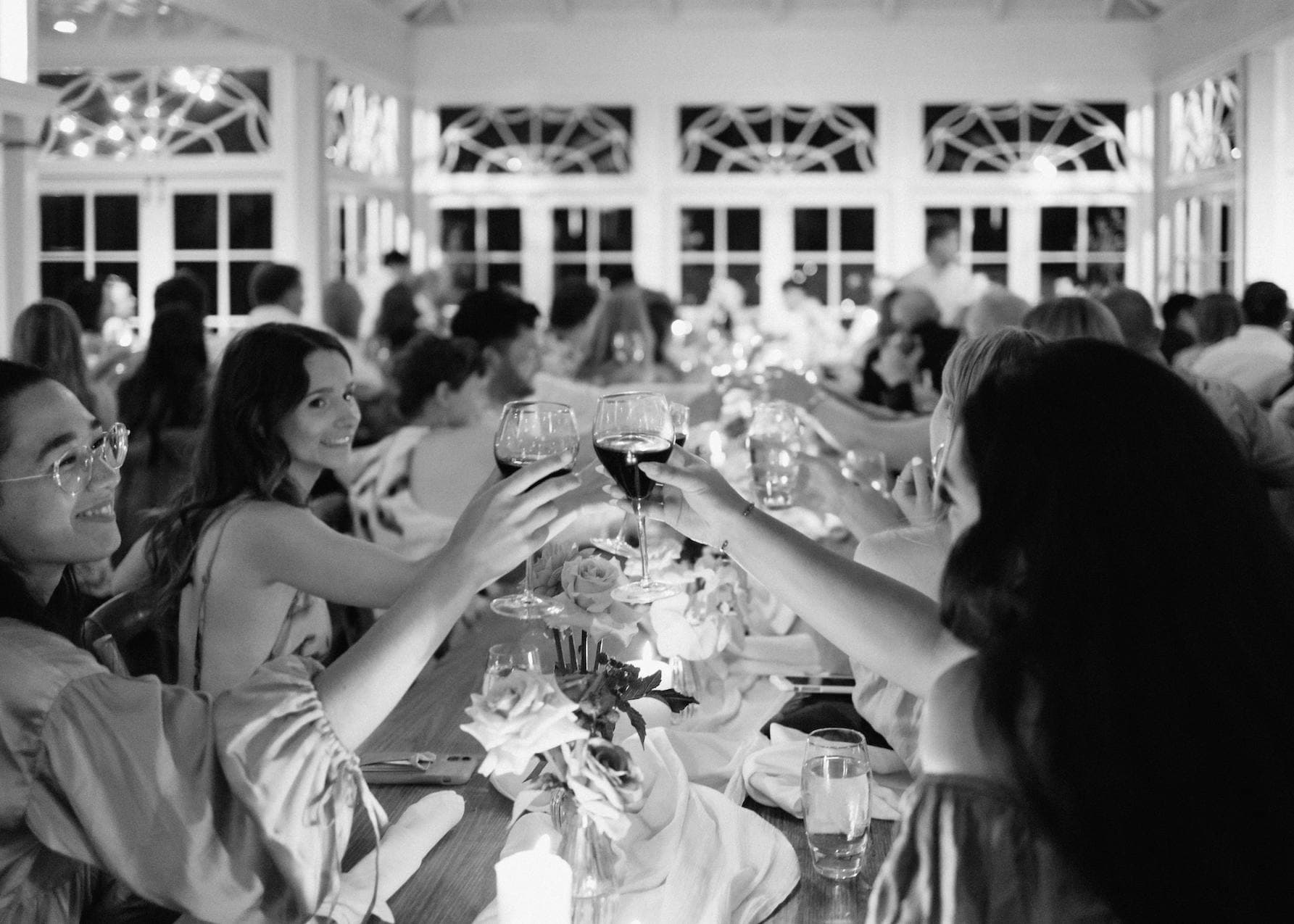 A group of people sit around a long, elegantly set dining table, raising their glasses in a toast. The room is well-lit with large windows and intricate decor. The atmosphere appears festive and celebratory.