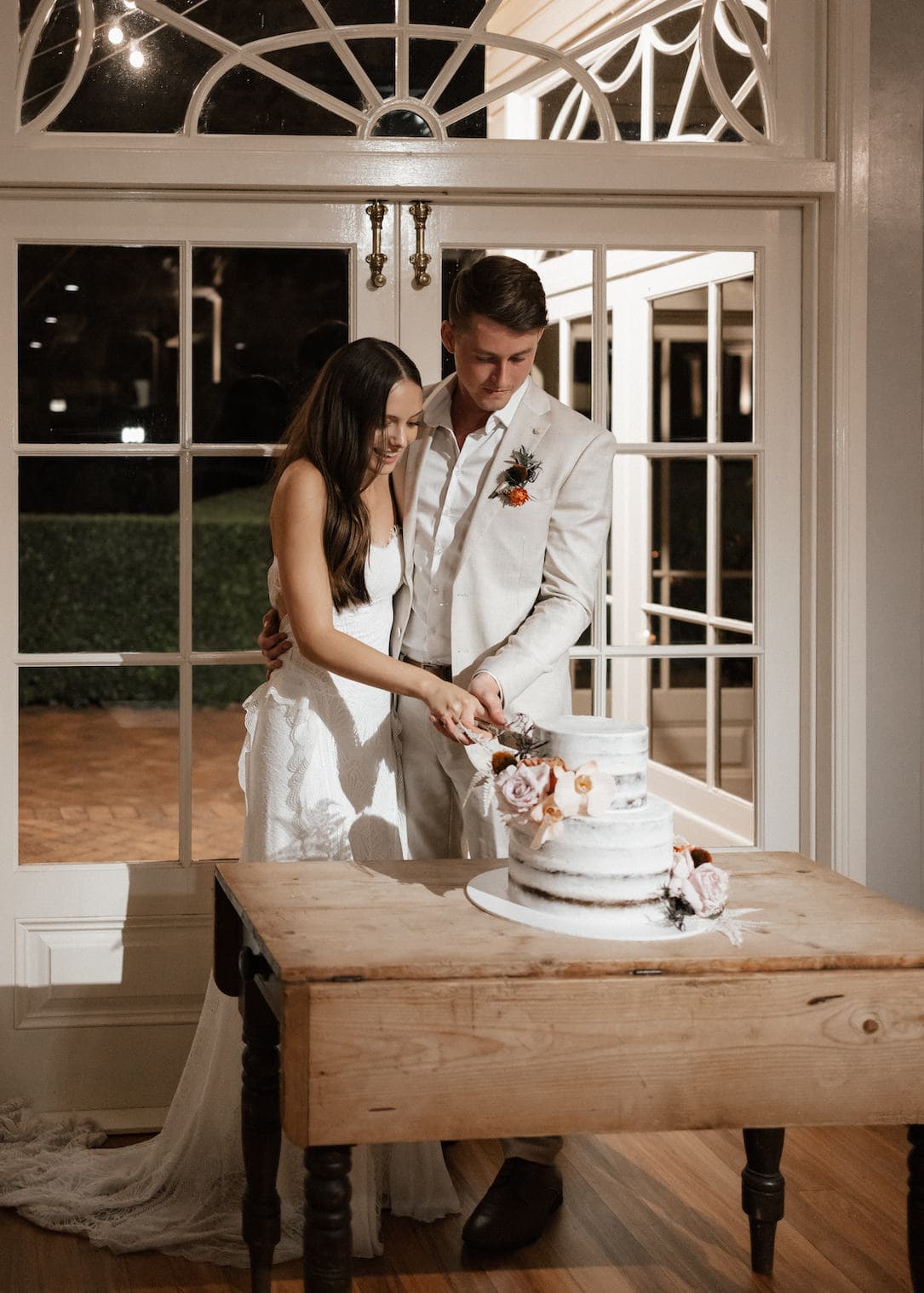 A couple in wedding attire stands together, cutting a tiered white cake decorated with pink flowers. They are in front of a large, ornate window, and the setting appears elegant and intimate. The bride wears a white dress, and the groom is in a light-colored suit.