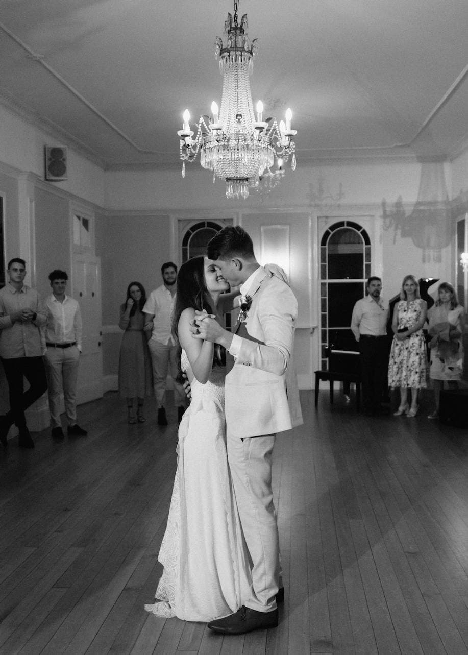 A couple shares a kiss during their first dance at a wedding, with the bride in a white gown and the groom in a light suit. They are under a chandelier in a spacious room with guests watching in the background. The photo is in black and white.