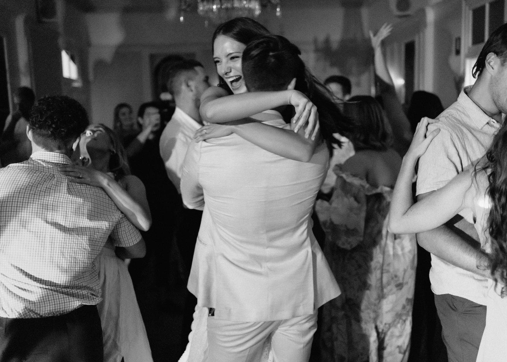 A black and white photo of a joyful couple embracing and dancing at a lively party or wedding reception. Around them, other couples are also dancing and enjoying the celebration. The atmosphere is filled with happiness and festivity.