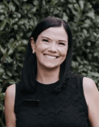 A woman with straight black hair smiles at the camera. She is wearing a sleeveless black top and small, round earrings. The background features green foliage.