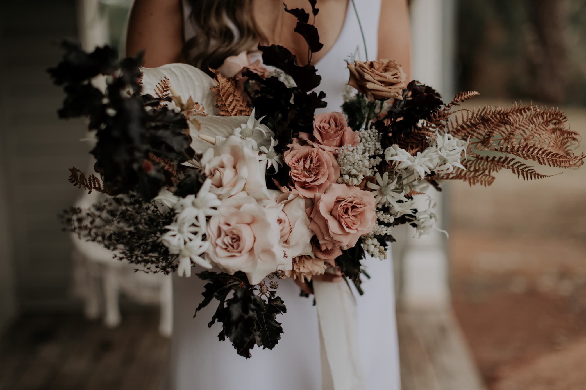 A person in a white dress is holding a large bouquet filled with pale pink roses, white flowers, and dark green foliage. The bouquet also includes dried brown ferns and leafy branches, creating a mix of soft and earthy tones.