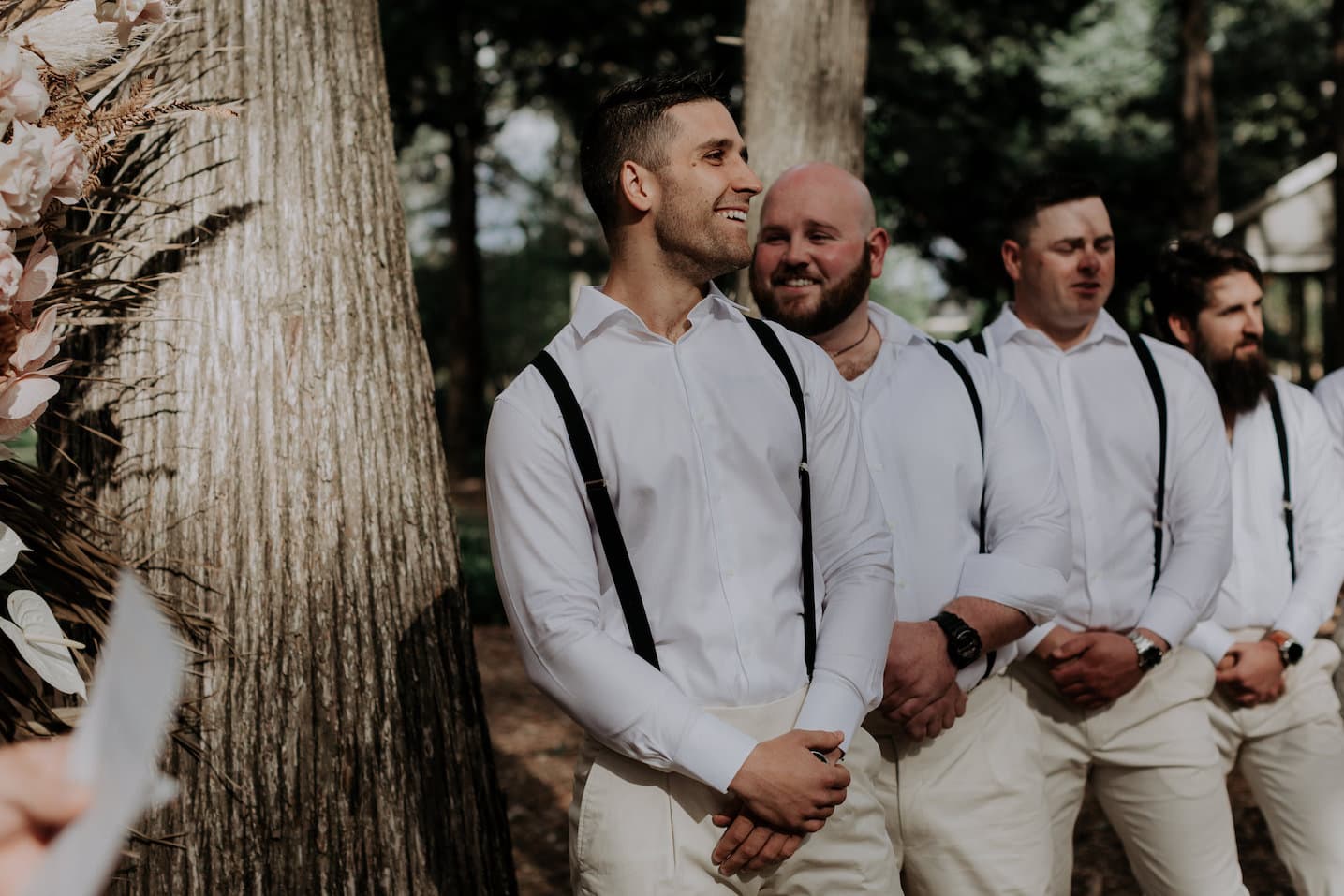 A group of four groomsmen stand in a line outdoors, all dressed in white shirts, black suspenders, and white pants. They look relaxed and happy. Tall trees are visible in the background, providing a natural and serene setting.