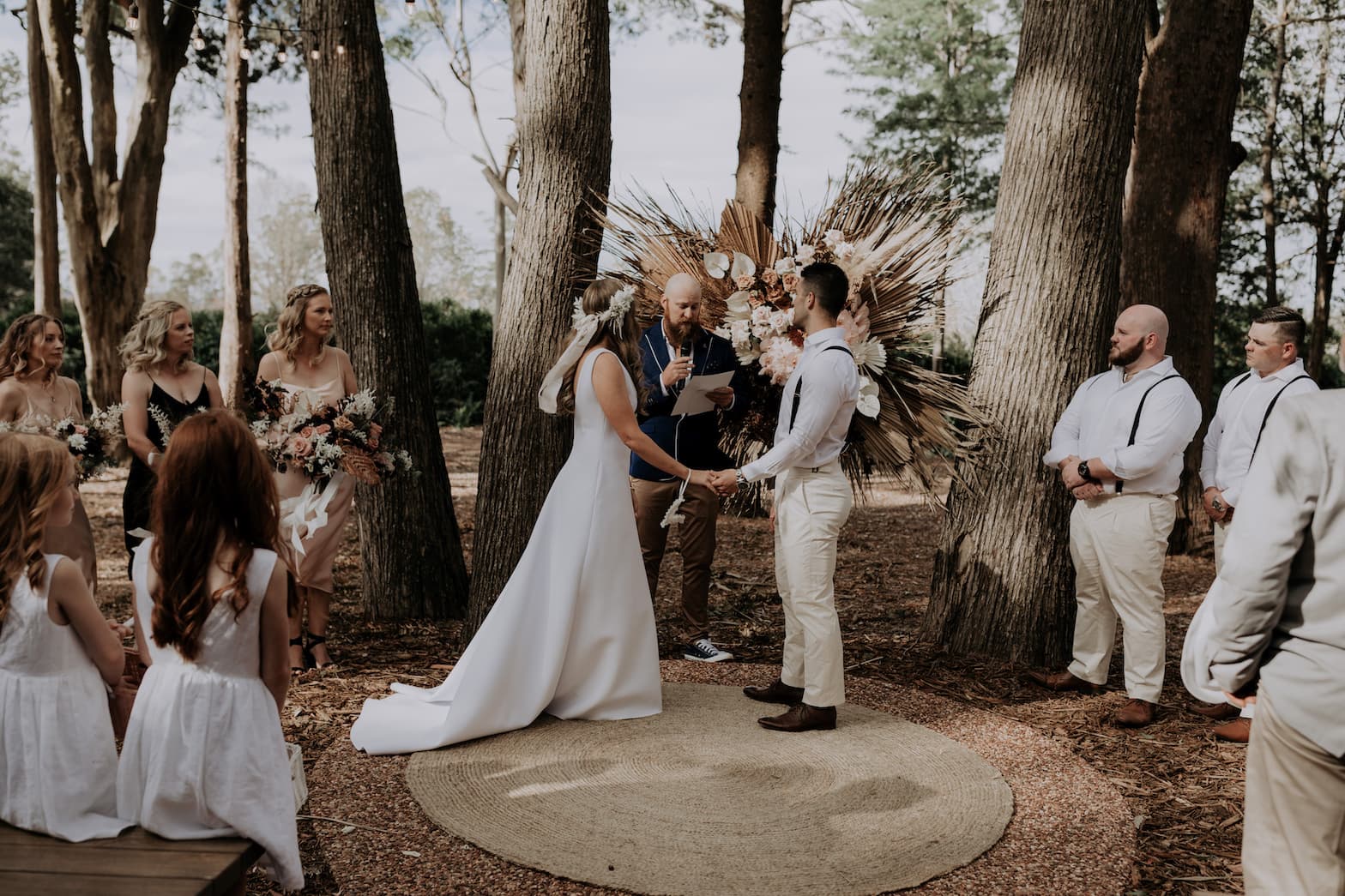 A couple stands holding hands during their outdoor wedding ceremony in a wooded area. The bride, in a white dress and floral headpiece, faces the groom in light-colored attire. Bridesmaids, a flower girl, and a groomsman look on. The officiant stands between the couple.