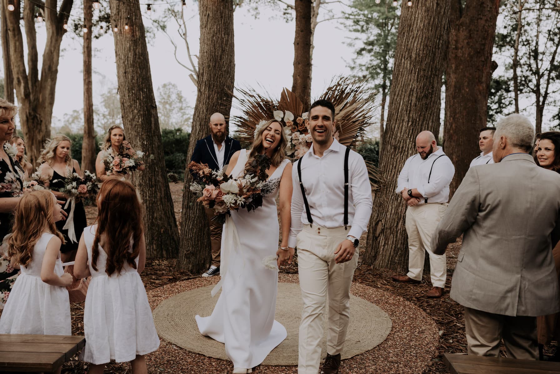 A joyful bride and groom walk hand-in-hand down an outdoor aisle, surrounded by smiling wedding guests in a forest setting. The bride holds a bouquet, and they both wear shades of white and beige. Flower girls stand nearby, holding small bouquets.