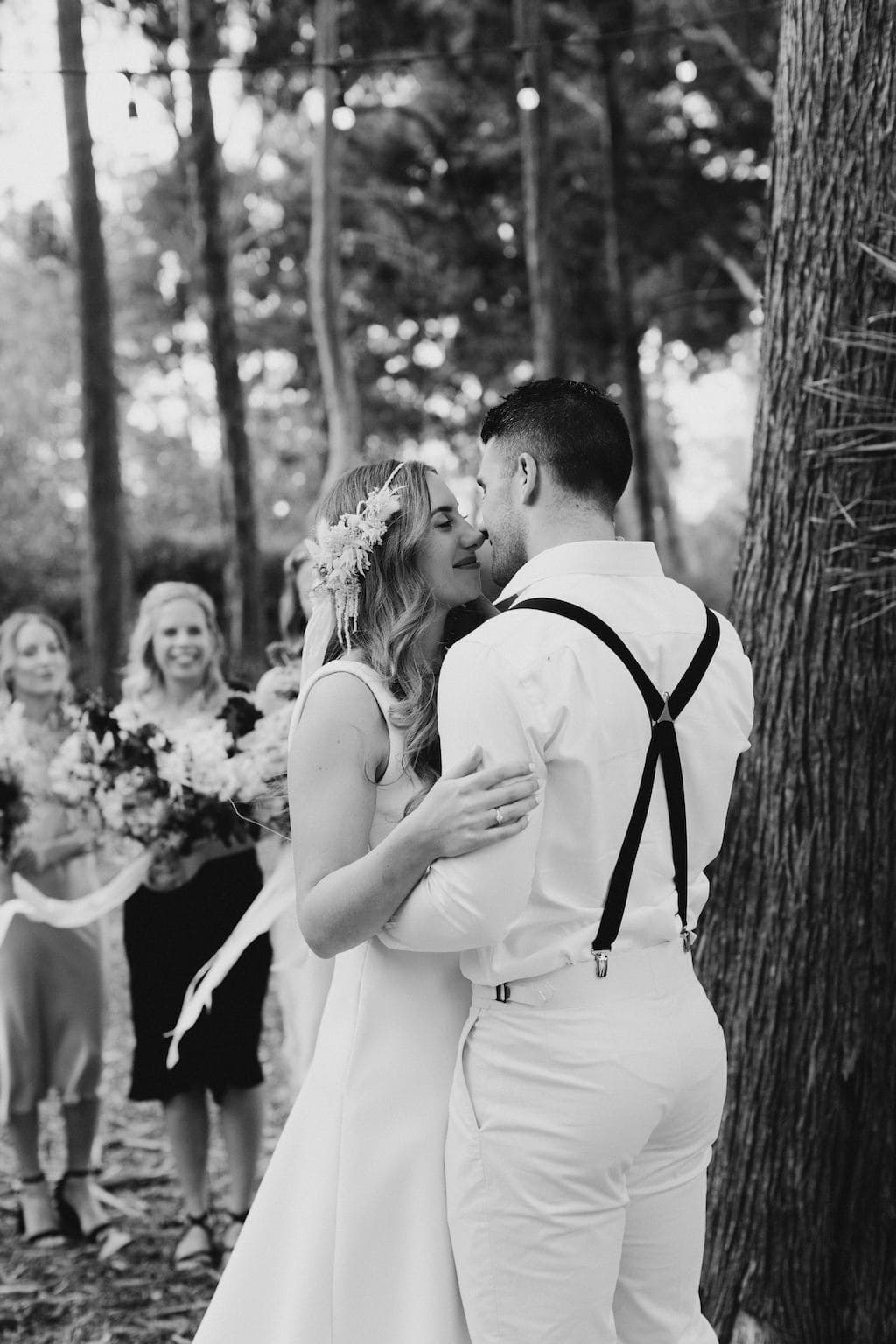 A newlywed couple shares an intimate dance in a forest. The bride, wearing a flower crown and dress, and the groom, in suspenders, gaze at each other lovingly. Bridesmaids with bouquets stand in the background, smiling. String lights hang above. Black and white.