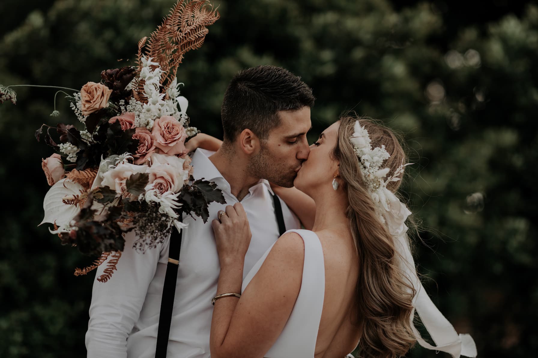 A bride and groom share a kiss outdoors, with the bride holding a large bouquet of pink roses, ferns, and other flowers. The bride wears a white dress and a floral headpiece, and the groom is dressed in a white shirt with suspenders. Green foliage backgrounds the scene.