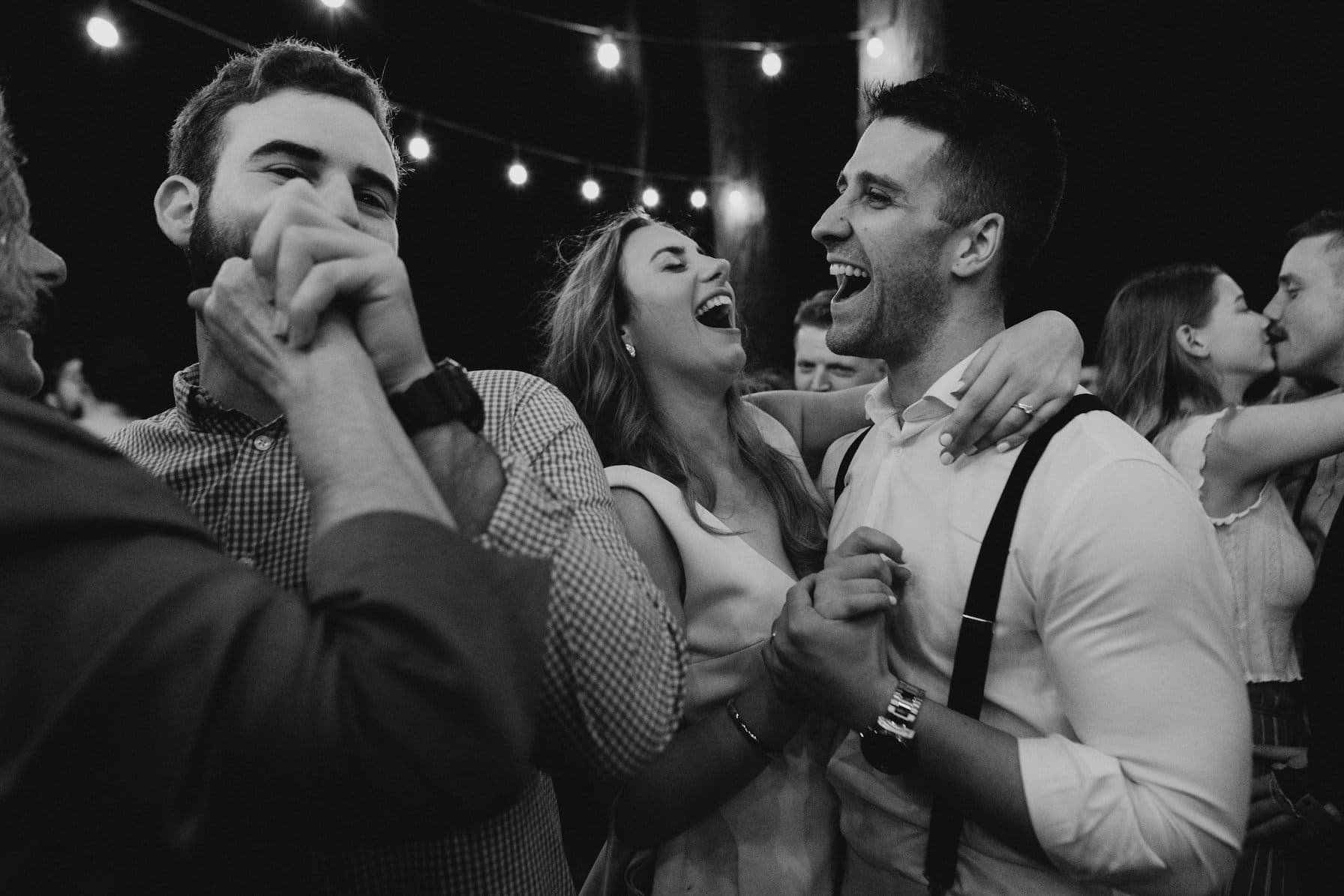 Black and white photo of people dancing and laughing at an outdoor event. String lights are hanging overhead. A man with a beard, a woman with long hair, and a man wearing suspenders are in the foreground, joyfully holding hands and smiling.