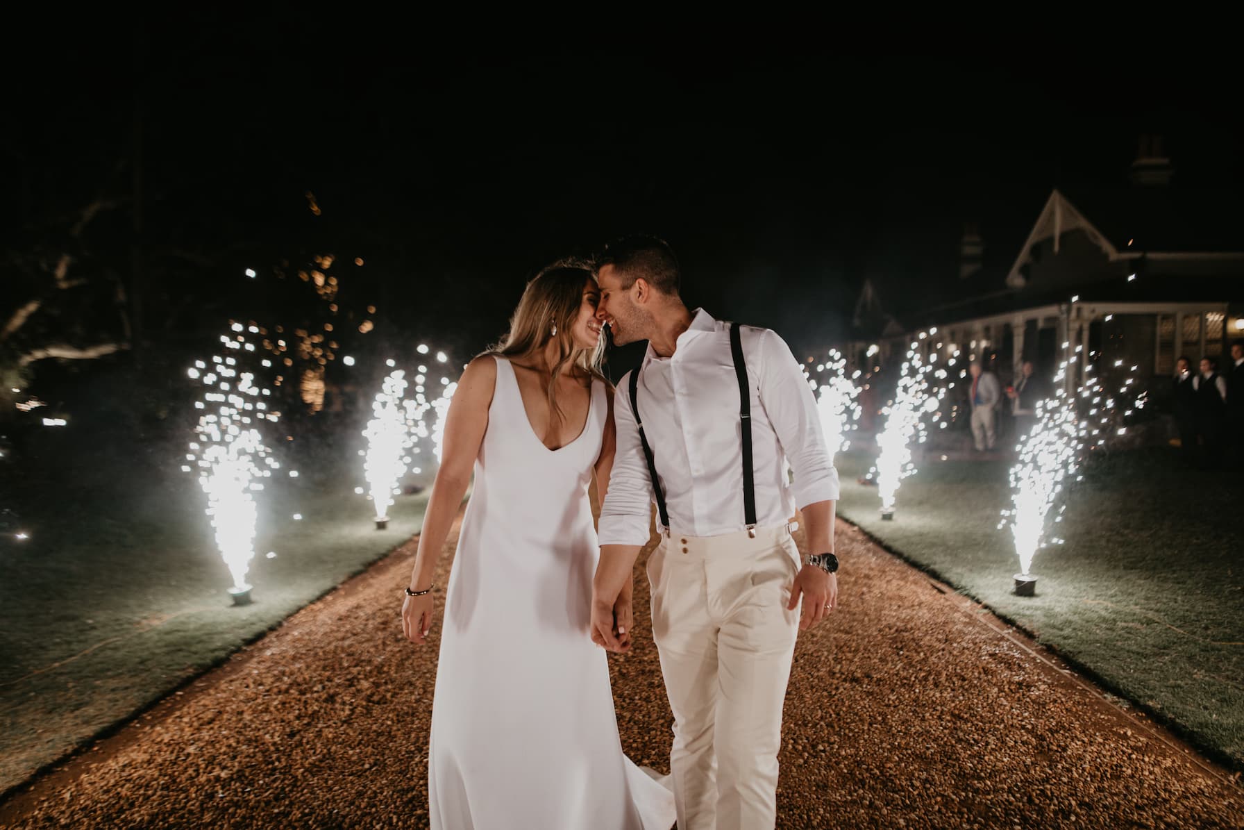 A couple walks hand in hand on a gravel path at night, dressed in white. The woman wears a sleeveless dress, and the man wears a shirt with suspenders. They lean towards each other, touching foreheads. Fireworks light the path, creating a festive atmosphere.