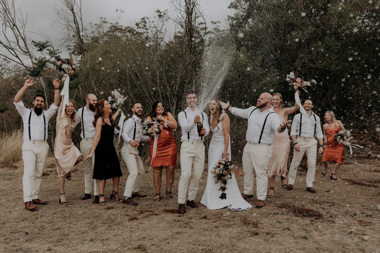 A joyful wedding party celebrates outdoors. The groom in a white shirt and beige pants pops a champagne bottle, spraying it into the air while the bride in a white dress holds a bouquet. Bridesmaids in pastel dresses and groomsmen in similar outfits cheer around them.