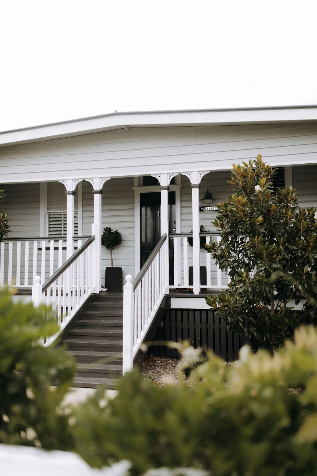 A charming white house features a covered front porch with decorative railings and trim. A set of stairs with a black handrail leads up to the front door. Lush greenery frames the pathway, adding a touch of nature to the inviting, well-maintained residence.