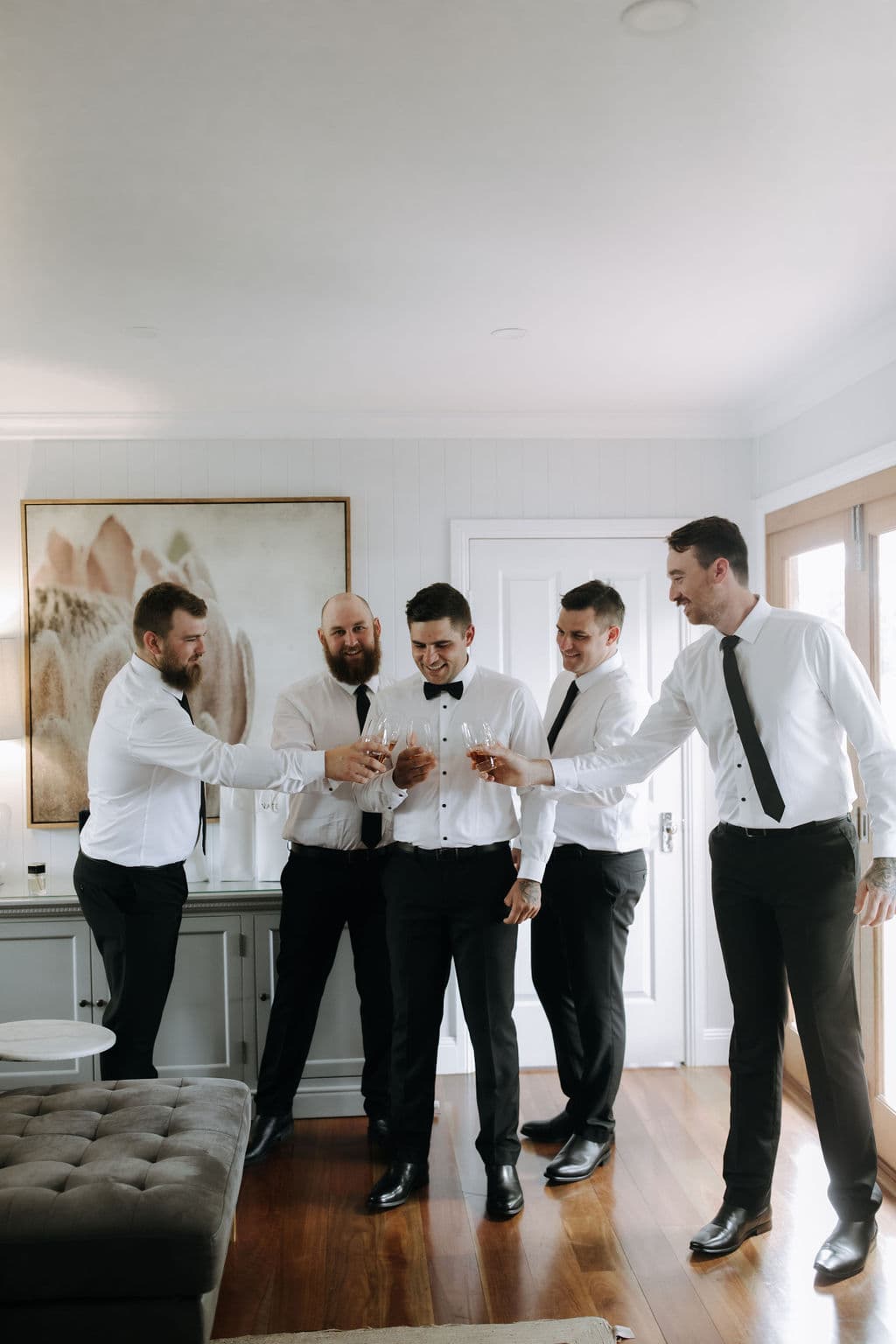 A group of five men dressed in white shirts and black pants are standing in a bright room, smiling and raising glasses in a toast. One man in the center appears to be the focus of the celebration, as the others surround him with joyful expressions.