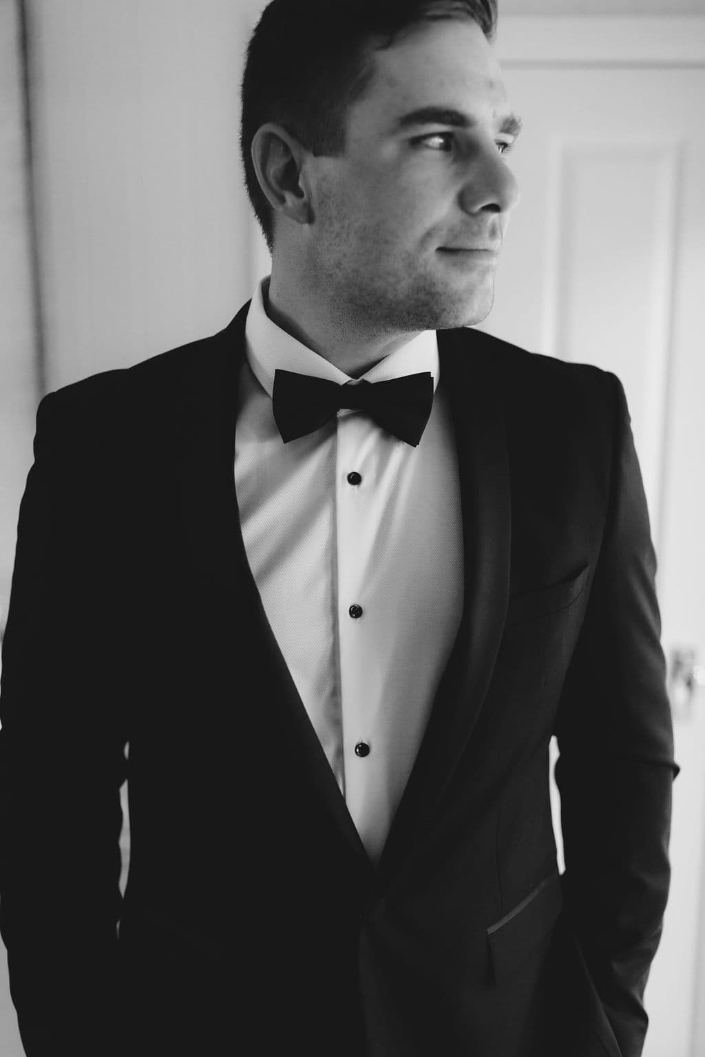 Black and white photo of a man in a formal black tuxedo with a bow tie. He is looking to the side with a serious expression, standing with his hands in his pockets. A door is visible in the background.