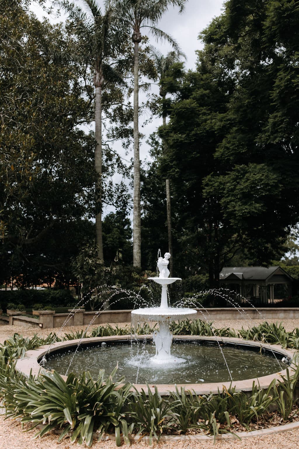 A white, two-tiered fountain with water cascading down, situated in a circular basin surrounded by lush green plants. Tall palm trees and dense foliage form the background under a cloudy sky.