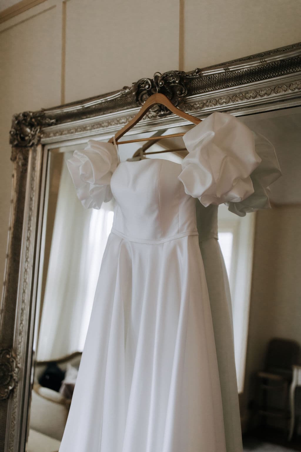 A white wedding dress with puffed sleeves hangs on a wooden hanger in front of a large ornate mirror. The elegant gown has a smooth, flowing design and is displayed in a softly lit room.