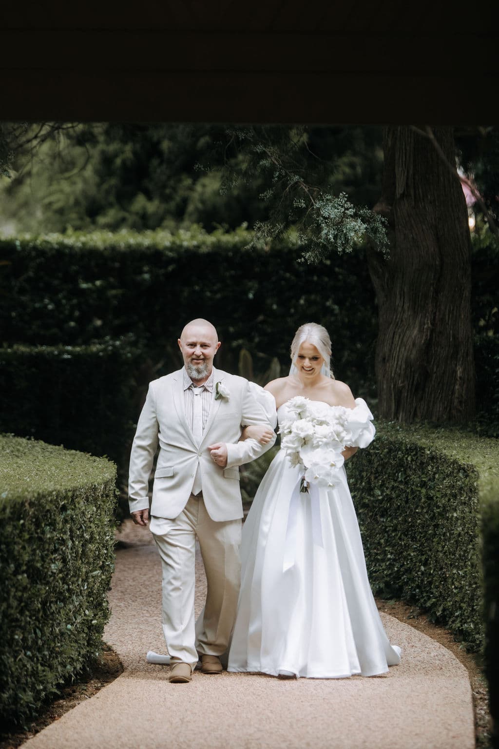 A bride in a white dress and holding a bouquet of white flowers walks arm in arm with an older man in a light-colored suit down a garden path lined with hedges. Both are smiling and walking under an archway.