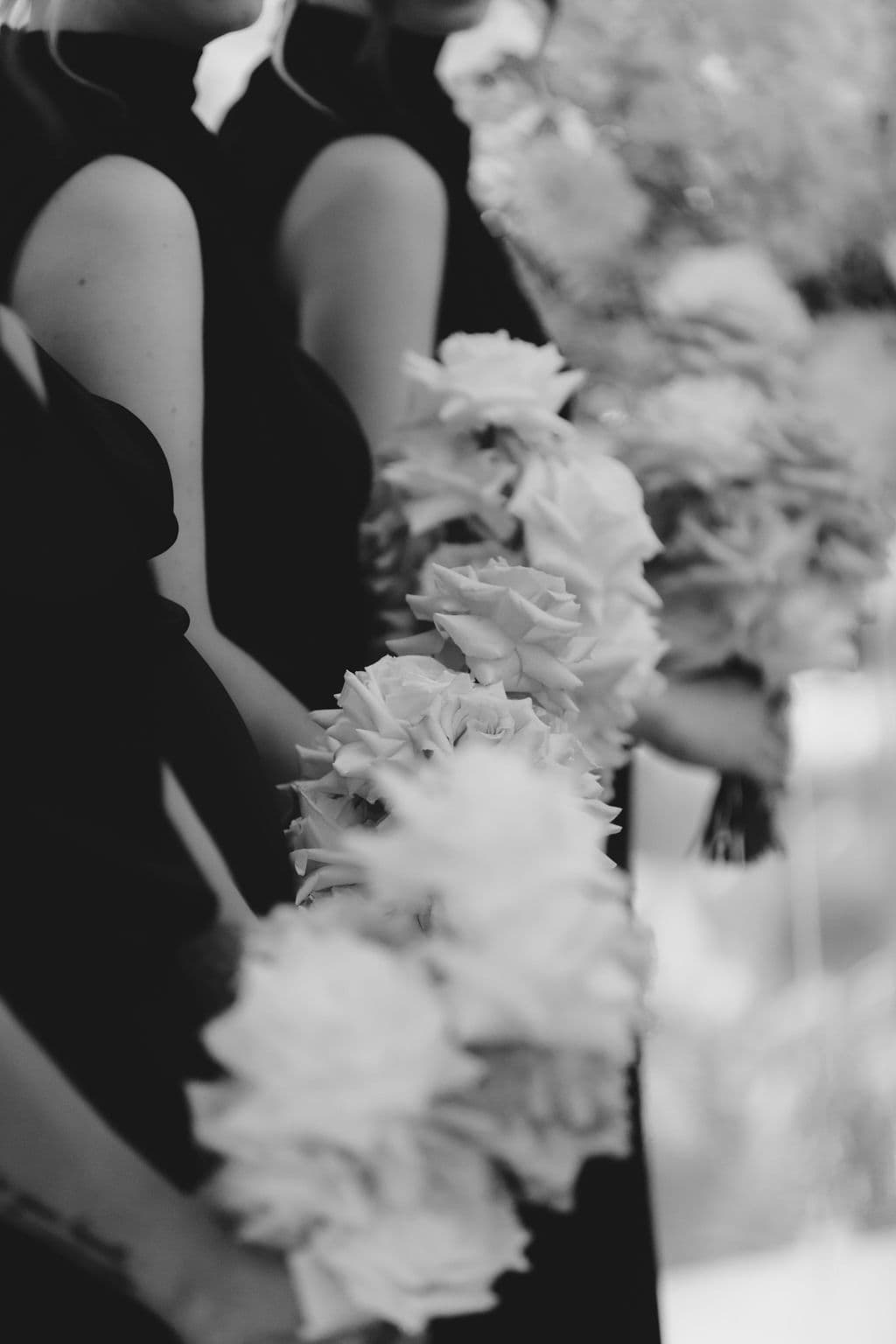 Black and white photo of three people standing side by side, each holding a bouquet of white flowers. The individuals are dressed in sleeveless, dark-colored attire, and the focus is on the floral arrangements they hold.