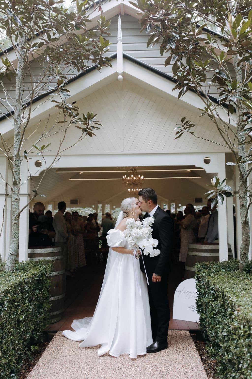 A bride and groom share a kiss in front of a charming white chapel surrounded by trees. The bride is wearing a flowing white gown and holding a bouquet of white flowers, while the groom is dressed in a black suit. Guests can be seen inside the chapel, celebrating.