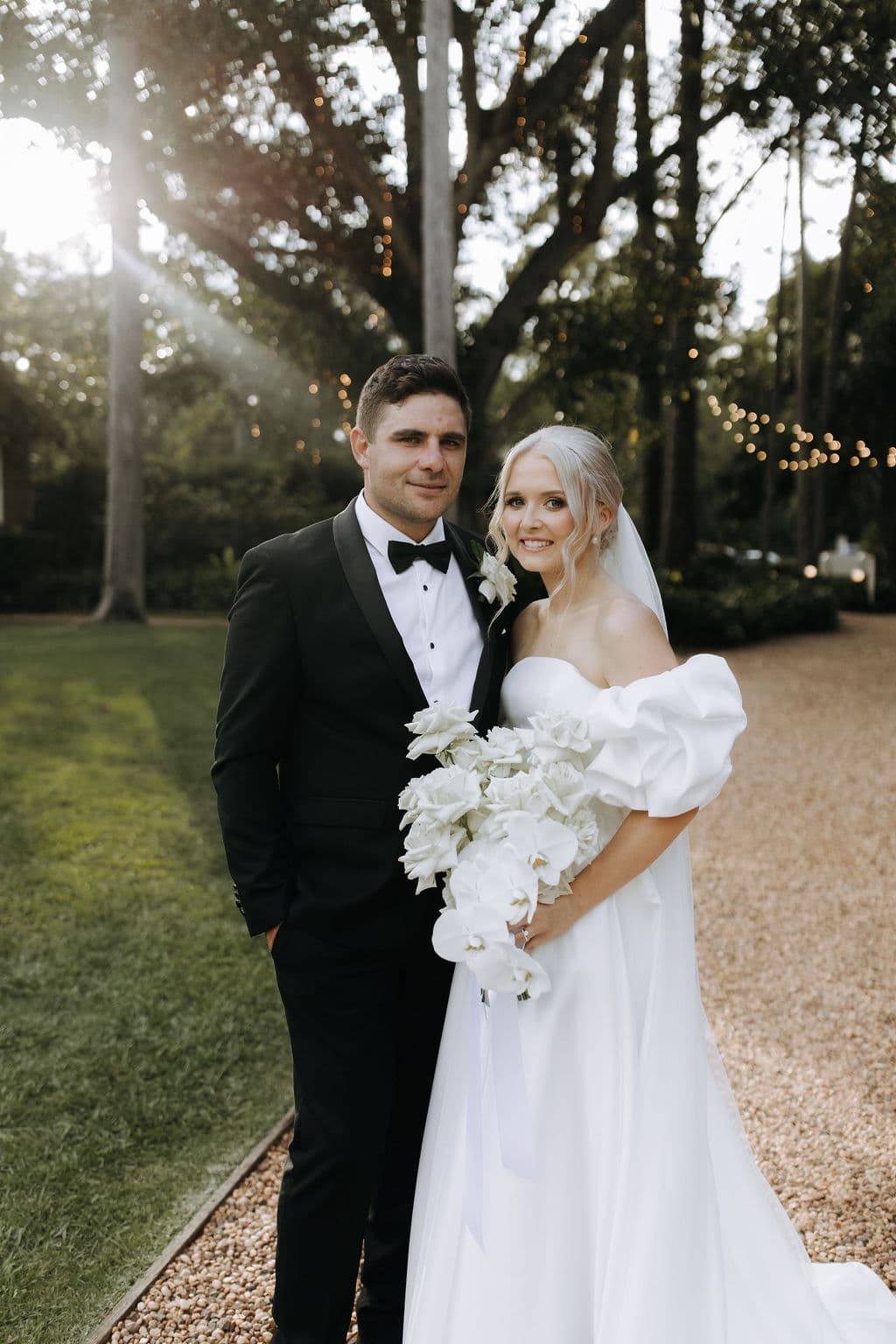 A couple dressed in wedding attire stands outdoors on a gravel pathway. The woman holds a bouquet of white flowers and wears a white off-shoulder wedding dress with a veil. The man wears a black tuxedo with a black bow tie. Trees and string lights are visible in the background.