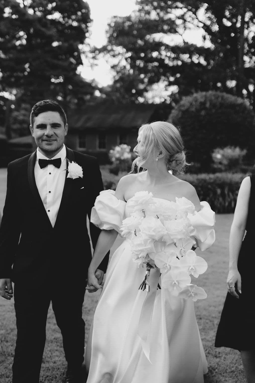 A couple dressed in formal wedding attire walks hand in hand outdoors. The man wears a black tuxedo with a bow tie, and the woman wears a white wedding gown with off-the-shoulder sleeves, carrying a bouquet of white flowers. Trees and a building are in the background.