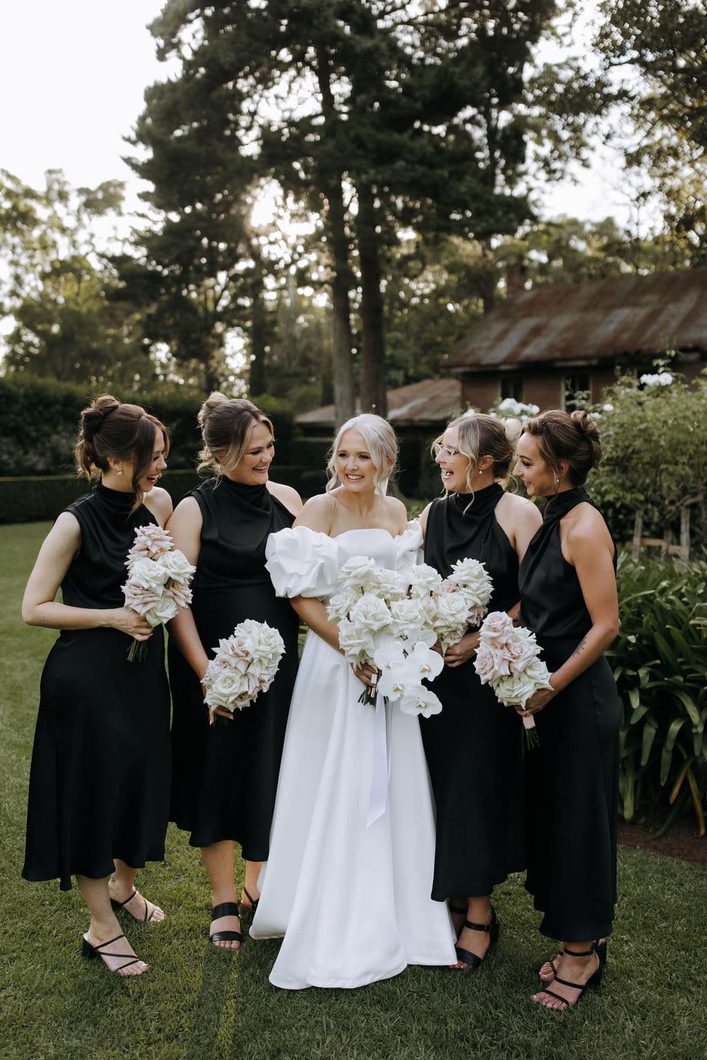 A bride in a white dress stands outdoors, surrounded by five bridesmaids in matching black dresses, all holding white bouquets. They are smiling and sharing a joyful moment in front of a lush, green background with tall trees and a rustic building.