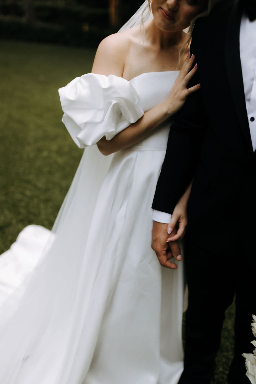 A bride in an elegant off-the-shoulder white gown stands next to a groom in a black tuxedo. She softly leans her head towards him and gently touches his arm. They hold hands, with a green grassy background behind them.