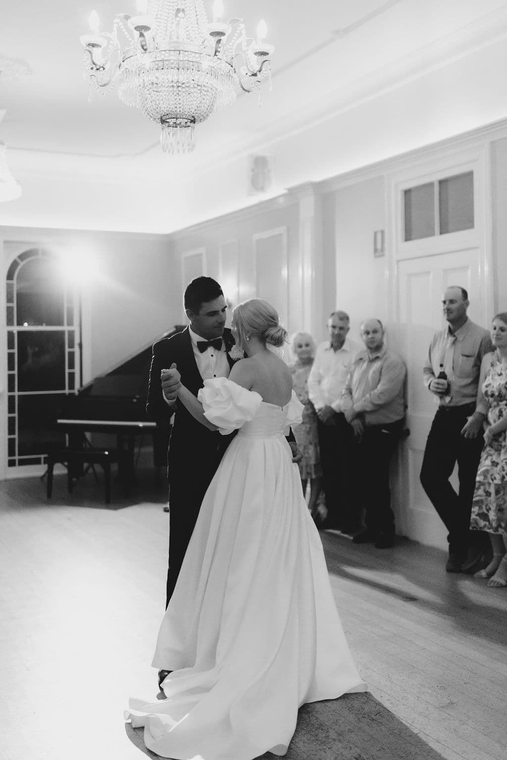 A bride and groom share their first dance in a spacious room with a chandelier overhead. The bride wears a long, flowing white gown, and the groom is in a black suit. Guests stand in the background watching the couple dance. There's a grand piano near the back wall.