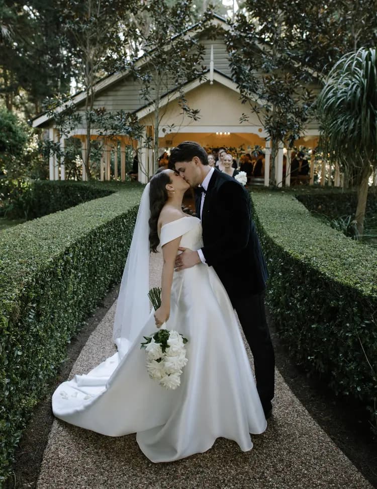 A couple dressed in wedding attire shares a kiss in front of a charming house. The bride, in an off-the-shoulder white gown with a long veil, holds a bouquet of white flowers. The groom, in a dark suit, embraces her on a gravel path bordered by lush green hedges.