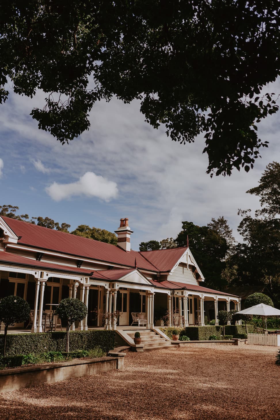 A grand house with a red roof stands amidst lush greenery. Its wraparound veranda is adorned with plants, while manicured gardens and a gravel driveway stretch out in front. The scene is framed by tree branches with a partly cloudy blue sky overhead.