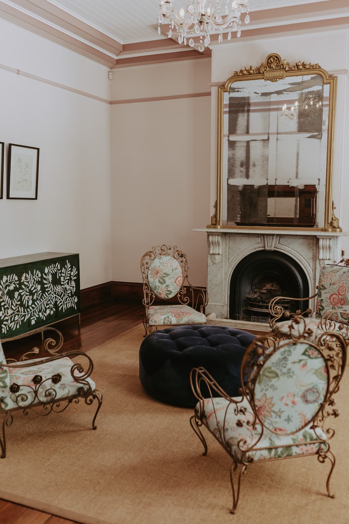 A cozy vintage-style living room with a chandelier, ornate mirror above a white fireplace, and floral-patterned wrought iron chairs surrounding a large blue tufted ottoman on a beige rug. Walls are pale pink with dark wooden accents.