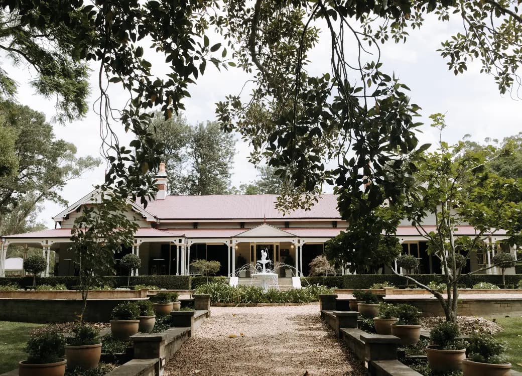 Image of a charming, single-story country house with a red roof, white trim, and a covered porch. It is surrounded by lush greenery, including large trees and potted plants, with a gravel pathway leading up to the entrance.