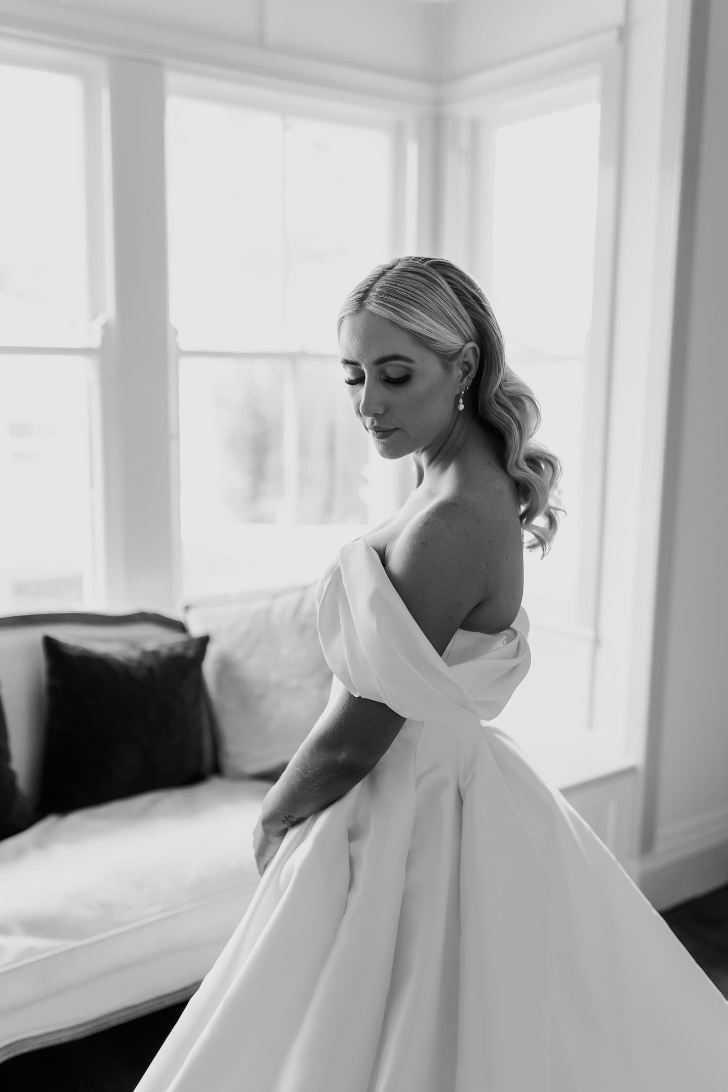 Black and white photo of a bride in an off-the-shoulder wedding gown, standing in front of a window. She has long, wavy hair and is looking down with a serene expression. Behind her, there is a cushioned bench with pillows.