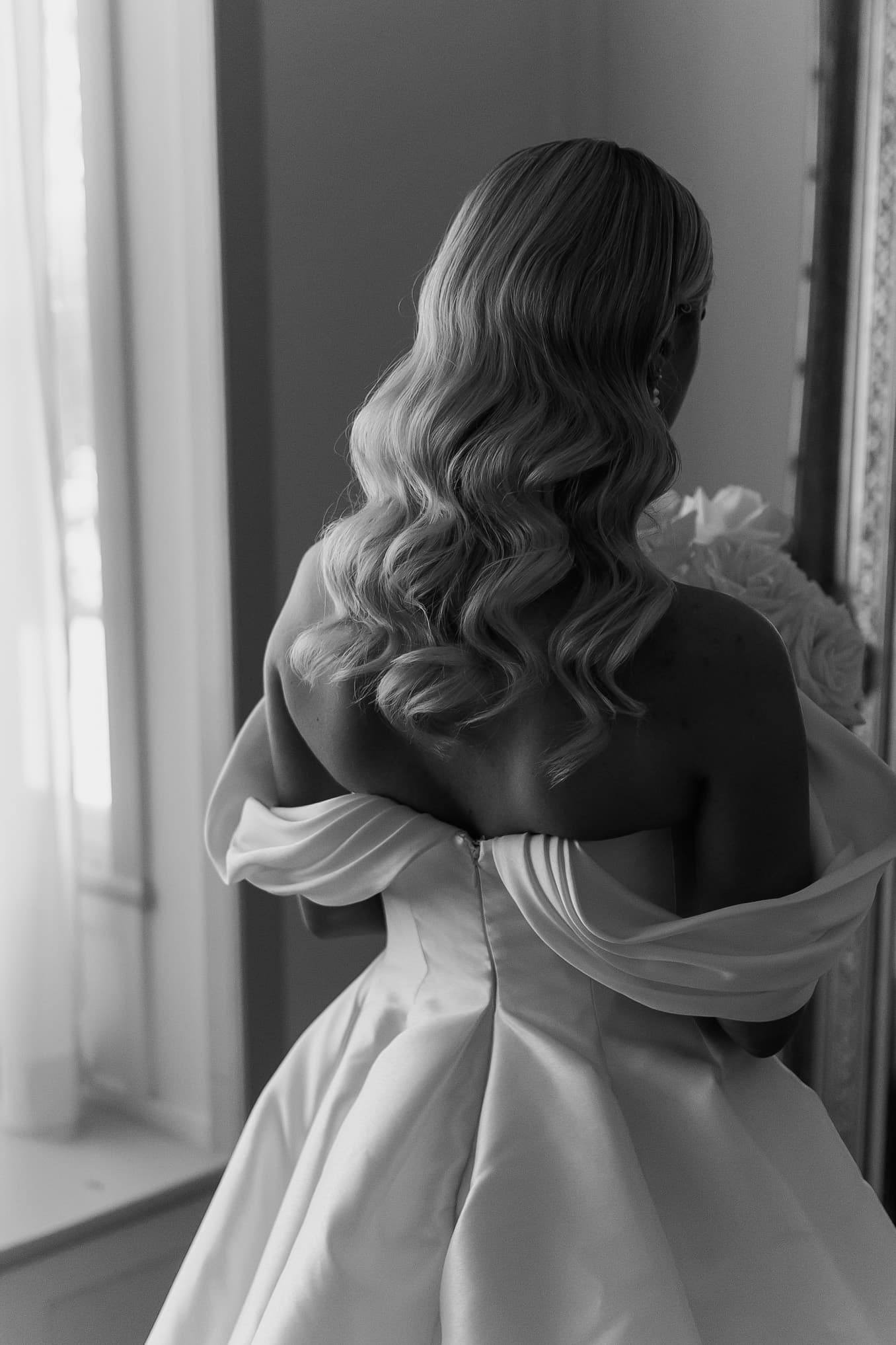 A woman in a white strapless gown with draped off-shoulder details stands near a window, holding a bouquet of flowers. Her long, wavy hair cascades down her back. The image is in black and white, capturing a serene and elegant moment.