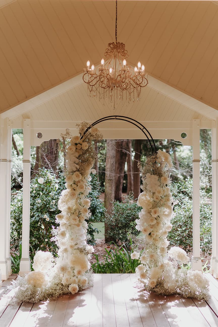 A decorated wedding arch stands under a covered pavilion adorned with white flowers and greenery. An elegant chandelier hangs from the ceiling above the arch. The scene is set against a background of lush trees and foliage, creating a serene, outdoor ambiance.