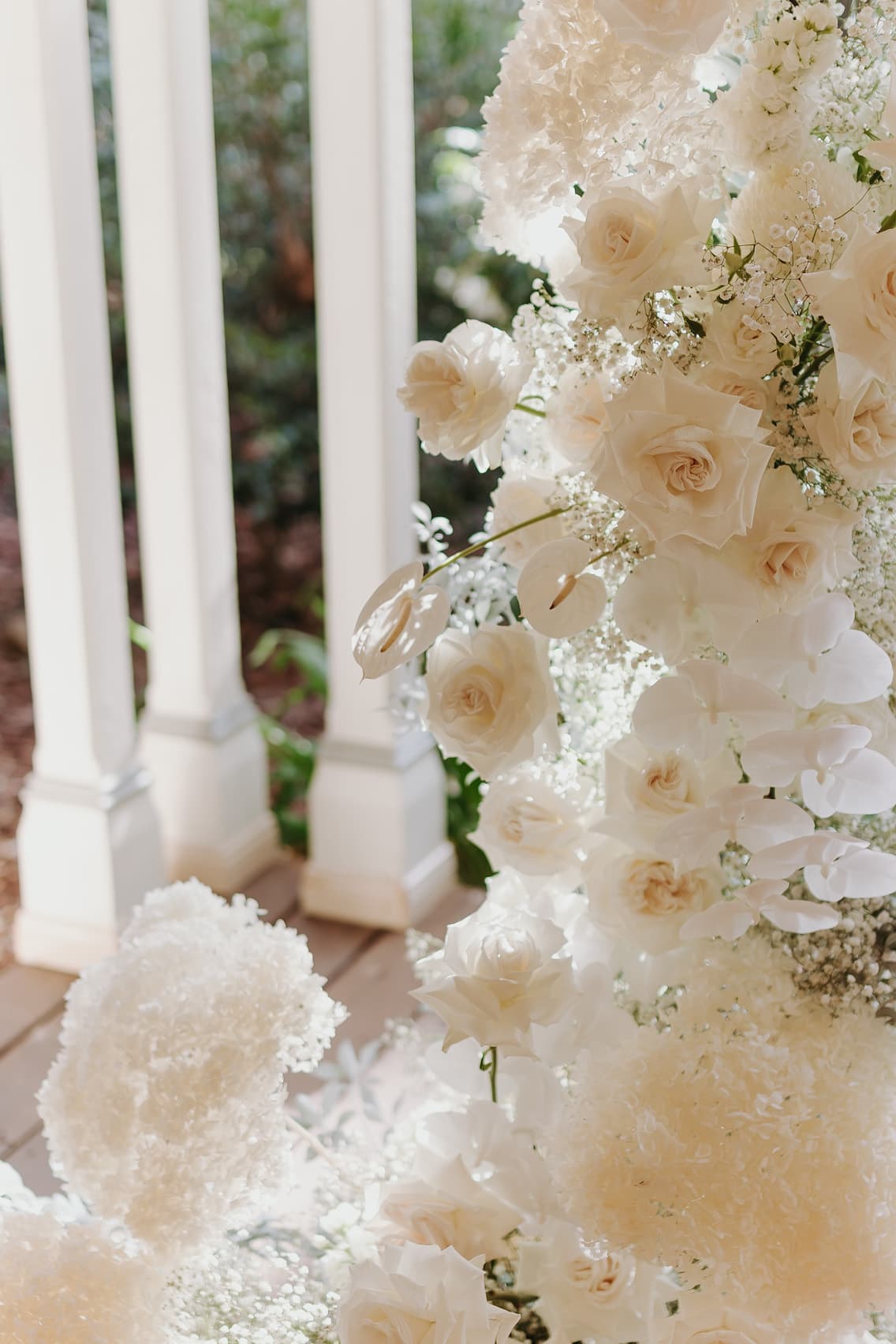 A close-up of a floral arrangement featuring a variety of white flowers, including roses and baby's breath. The arrangement is set against the backdrop of a white porch railing with greenery in the background. The overall appearance is elegant and serene.