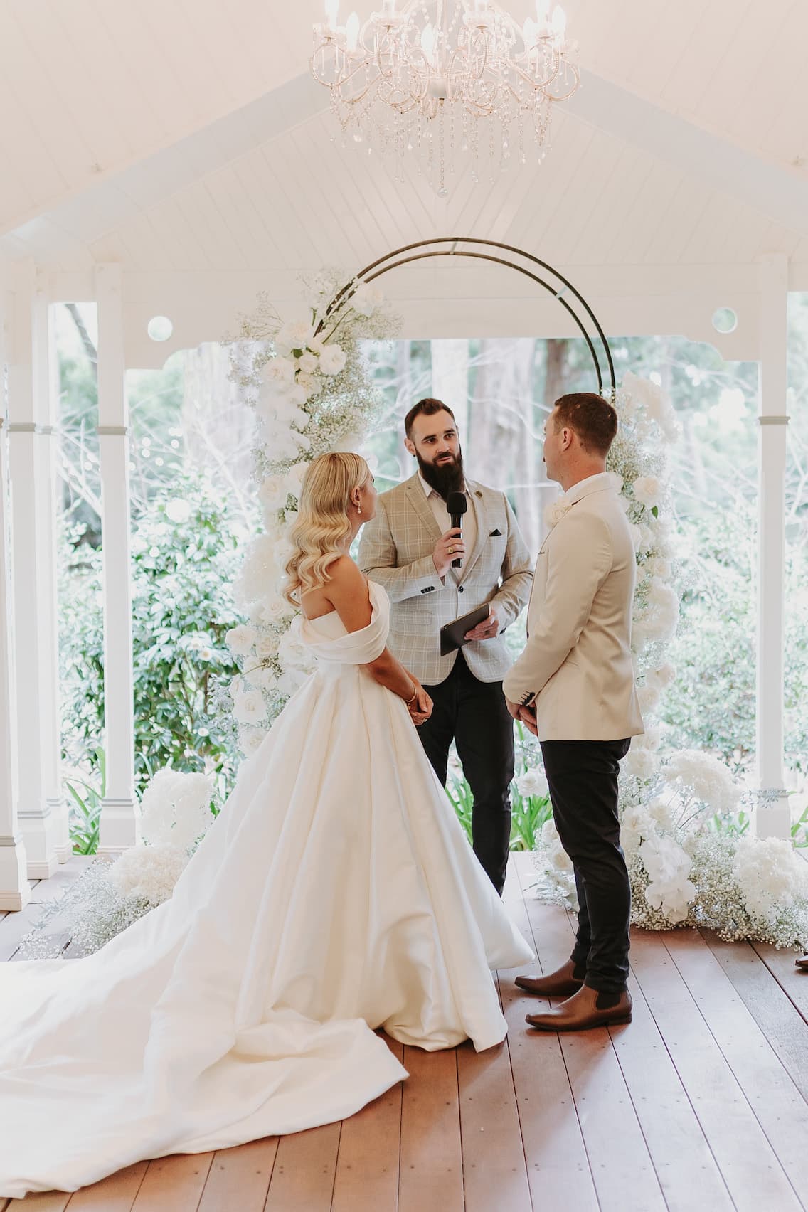 A bride and groom stand facing each other under a floral arch during their wedding ceremony. The bride is in a white gown with a long train, and the groom is in a light-colored jacket and dark pants. The officiant, holding a microphone, is speaking to them.