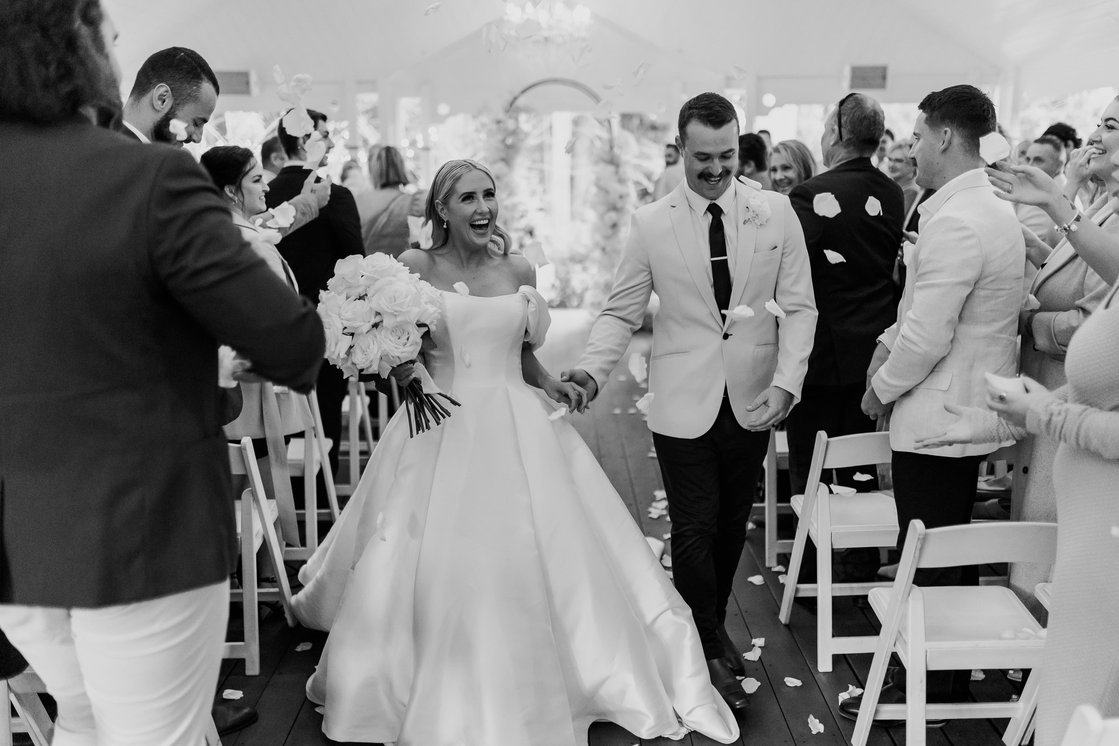 A bride and groom walk hand-in-hand down the aisle, both smiling joyfully. The bride carries a bouquet of white flowers and wears a flowing white gown, while the groom is dressed in a light-colored suit. Guests on either side throw rose petals, celebrating their union.