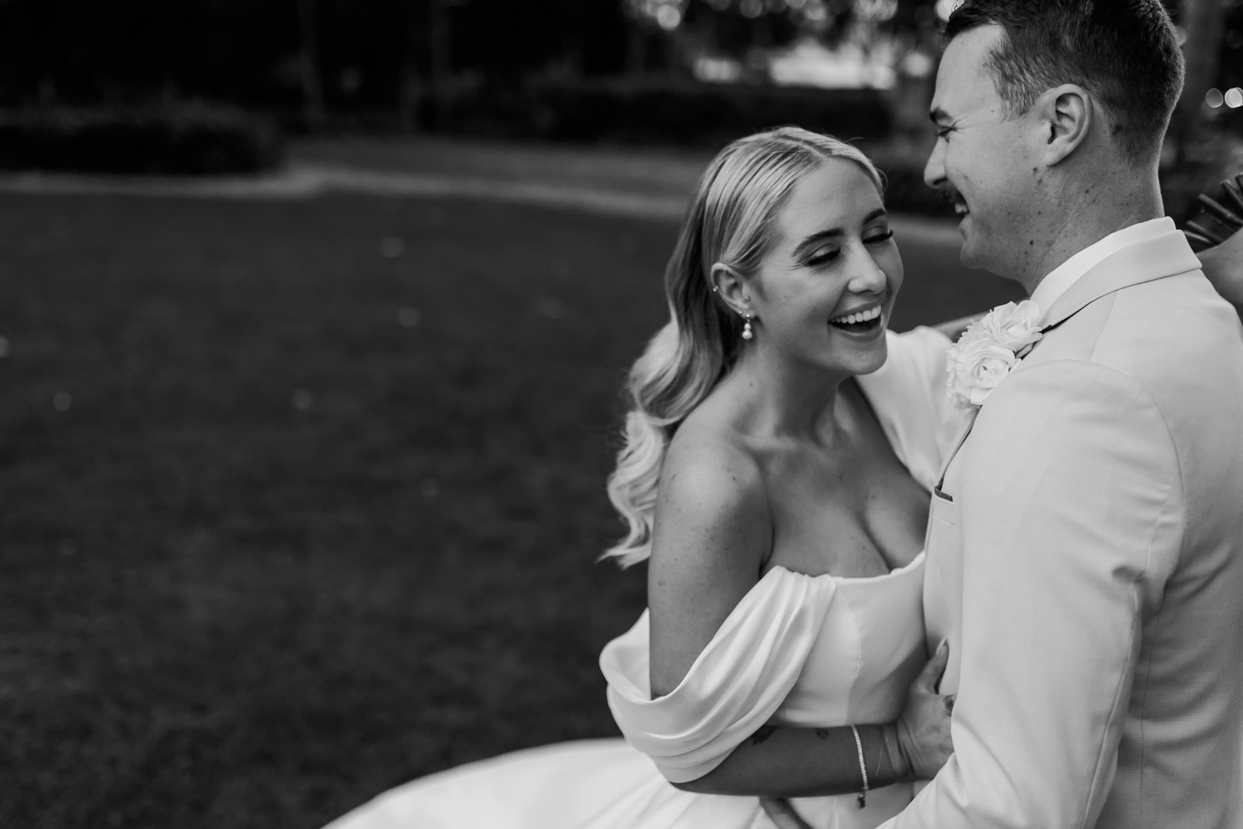 A black-and-white photo of a bride and groom on their wedding day. The bride, with long wavy hair and an off-the-shoulder dress, smiles while looking at the groom, who is wearing a suit. They are standing outside on a lawn, sharing a joyful moment.