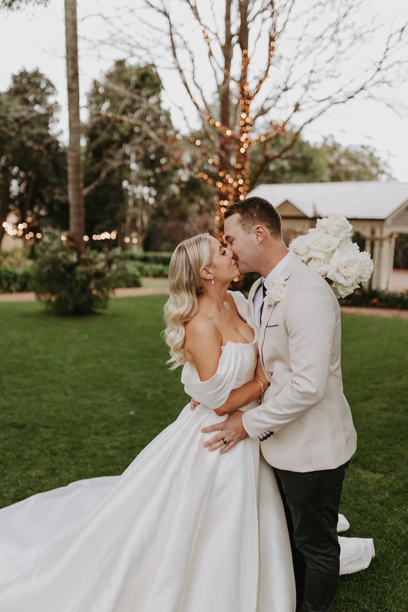 A bride and groom share a kiss on a lawn in front of a tree wrapped with lights. The bride is wearing an off-shoulder white wedding dress and holding a bouquet of white flowers, while the groom is dressed in a light-colored suit.
