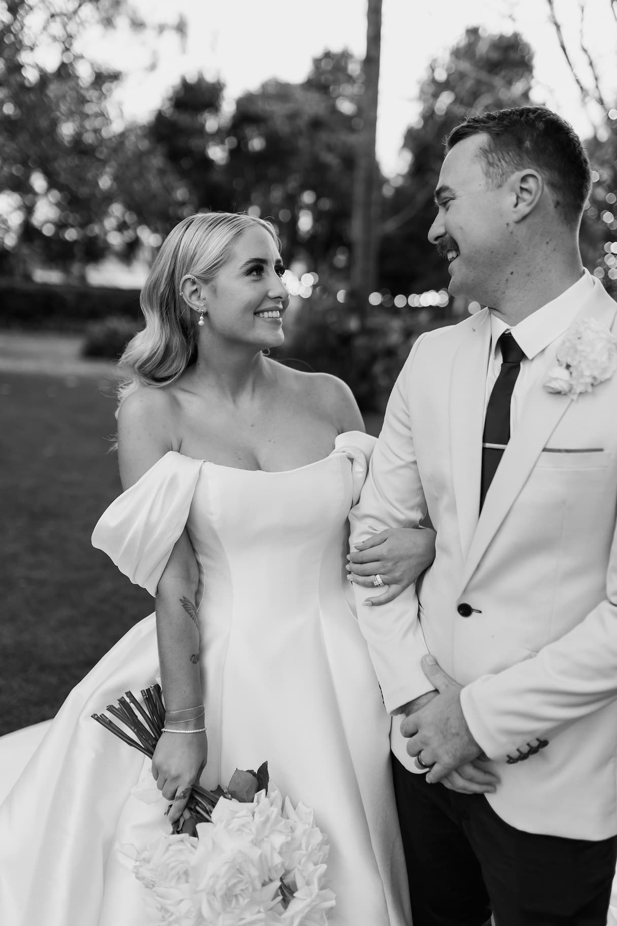 A black and white photo of a bride and groom on their wedding day. The bride is wearing an off-the-shoulder gown and holding a bouquet, while the groom is dressed in a light-colored suit. They are standing outdoors, looking at each other and smiling.