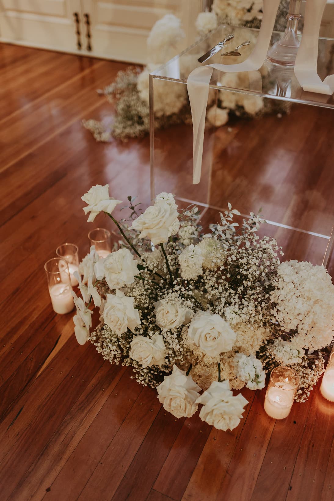 A wedding decoration featuring white roses and baby's breath arranged at the base of a clear acrylic stand. Several lit candles are placed around the flowers, casting a warm glow on the polished wooden floor. Elegant white fabric drapes from the stand above.