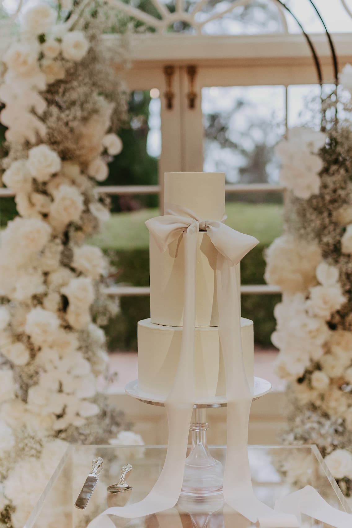 A three-tiered white wedding cake adorned with a large white ribbon stands elegantly on a clear pedestal. The backdrop features lush white floral arrangements and a window through which greenery is visible. Two silver serving utensils lie on a transparent surface nearby.