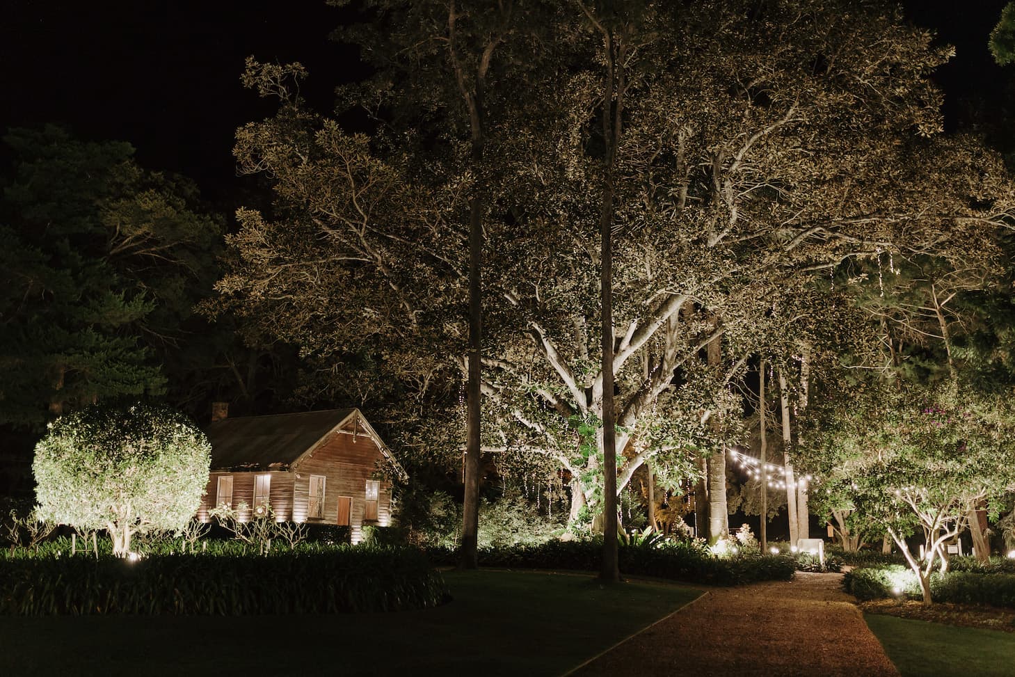 A quaint wooden cabin is nestled among tall trees and illuminated by soft, warm lights at night. String lights are visible to the right, adding a cozy ambiance to the serene, secluded forest setting. A gravel path leads towards the cabin.